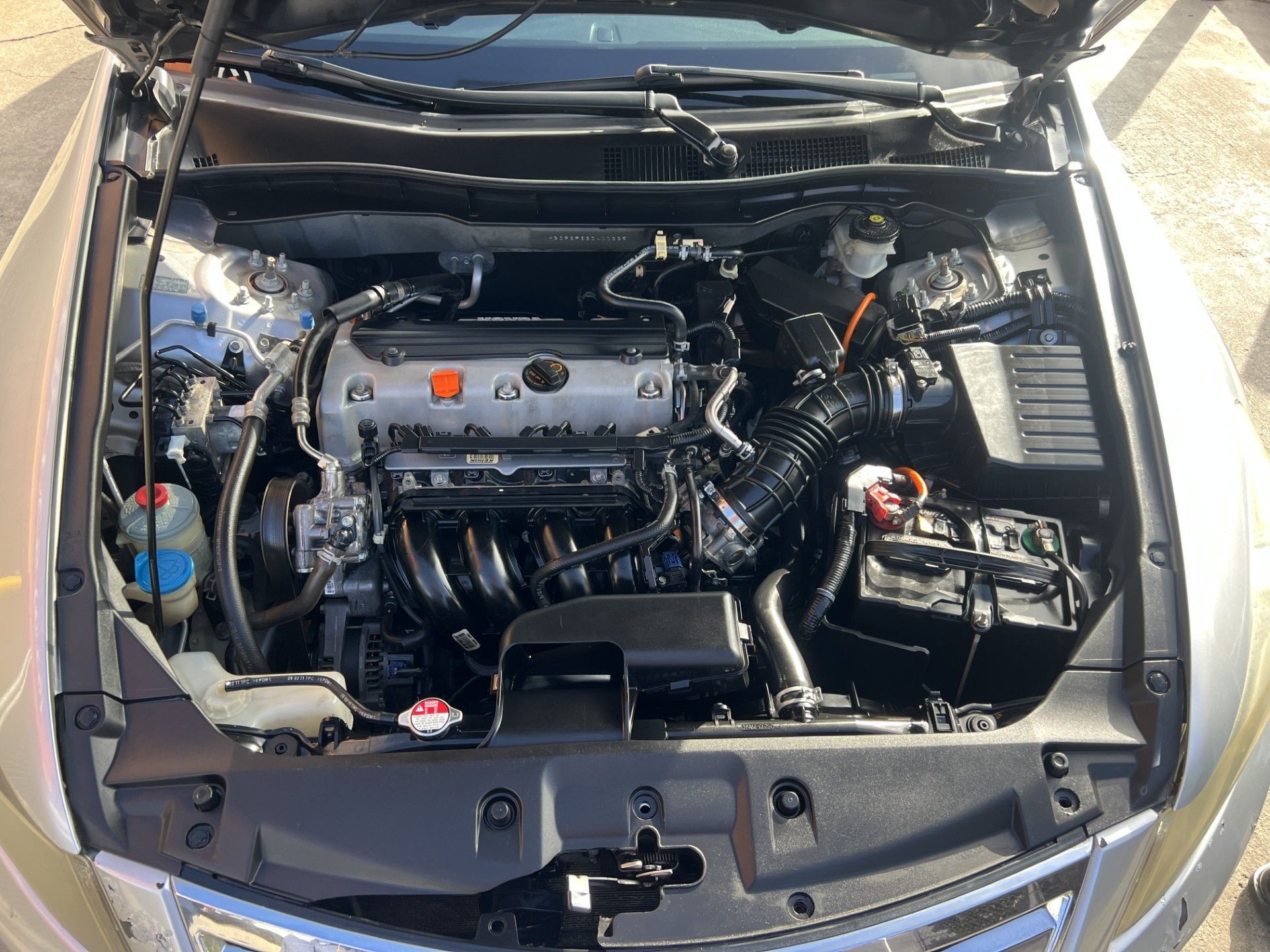 An open engine bay of a silver sedan, featuring a central engine, black air intake, battery, and fluid reservoirs.