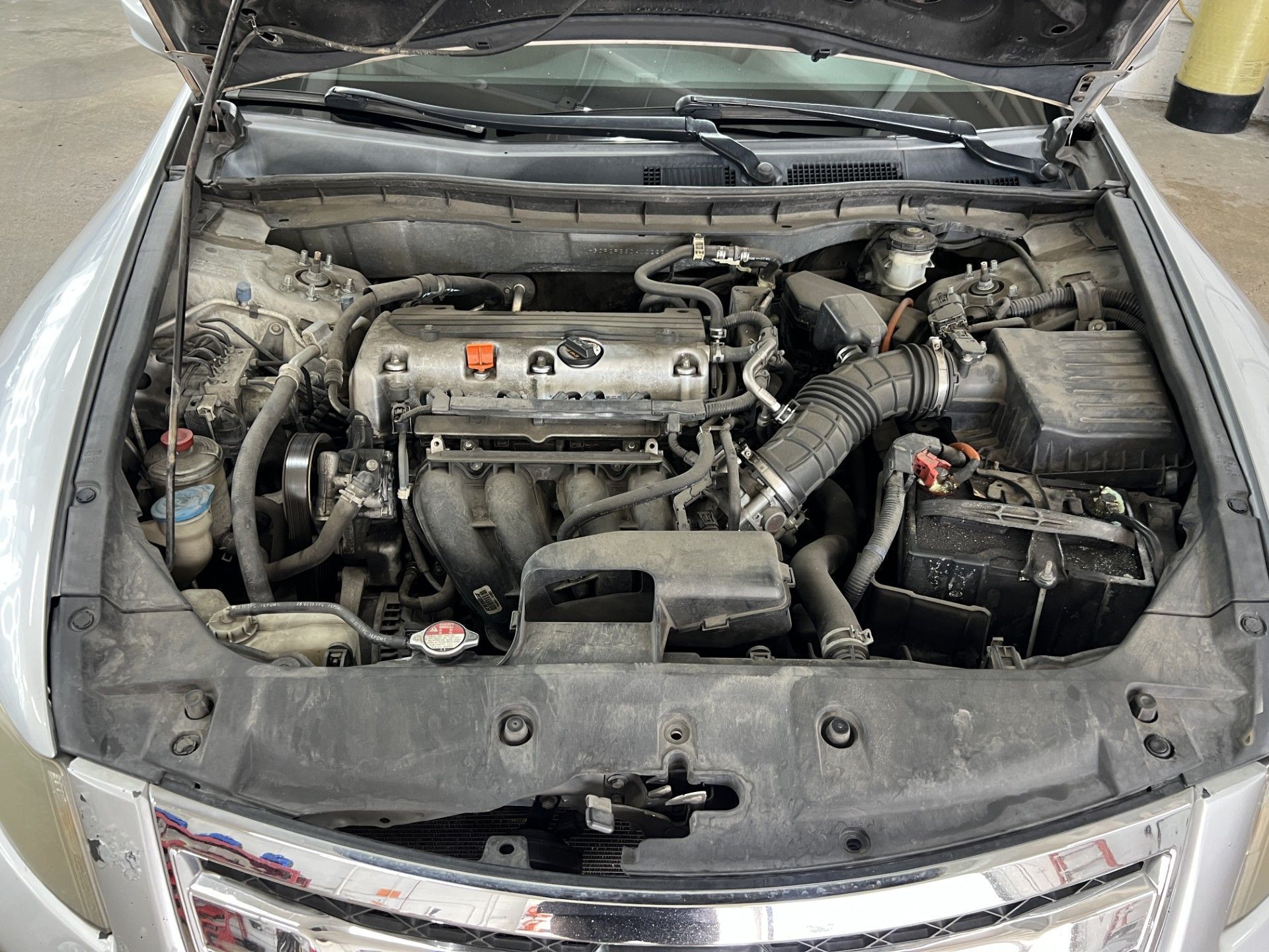 A view of an open car hood showing a dusty engine bay with the engine, battery, and intake components visible.