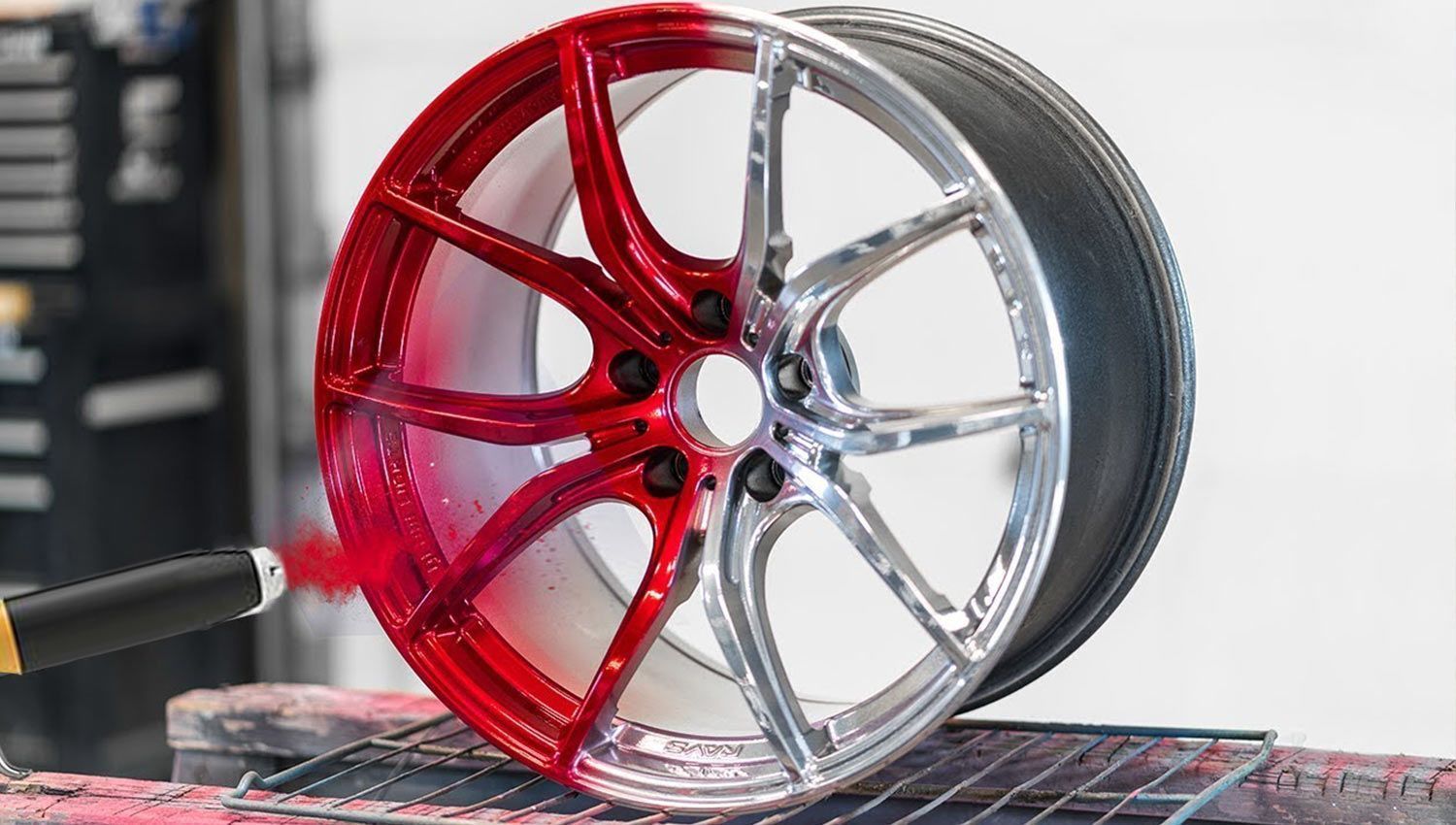 A close-up view of a metal car wheel being painted with a bright red spray, transitioning from silver to red.