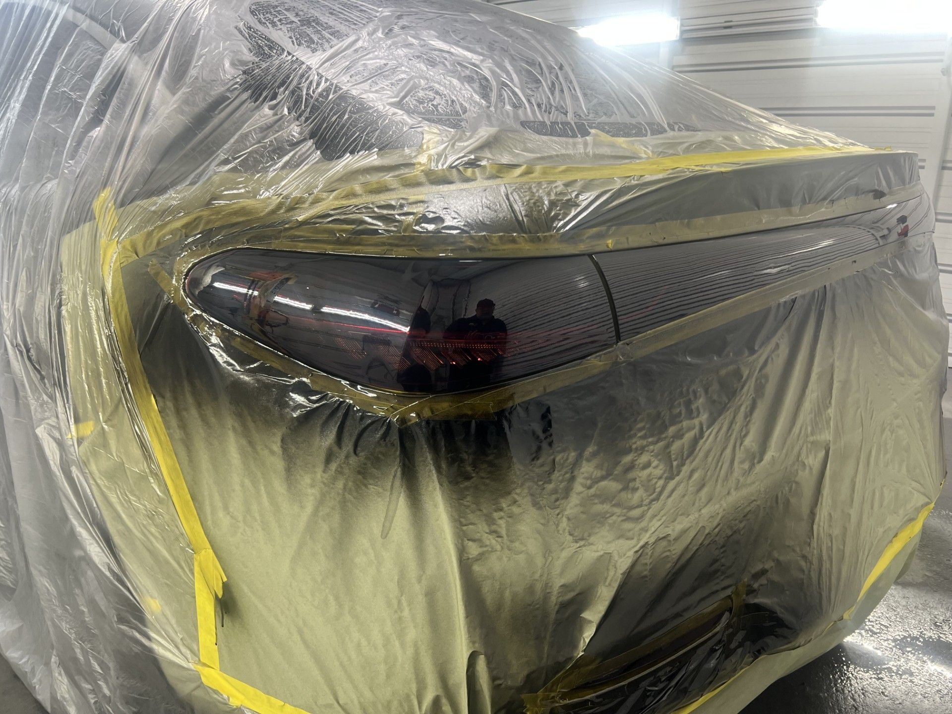 A vehicle's taillight covered in plastic film and masking tape, prepared for a professional paint or tint application.