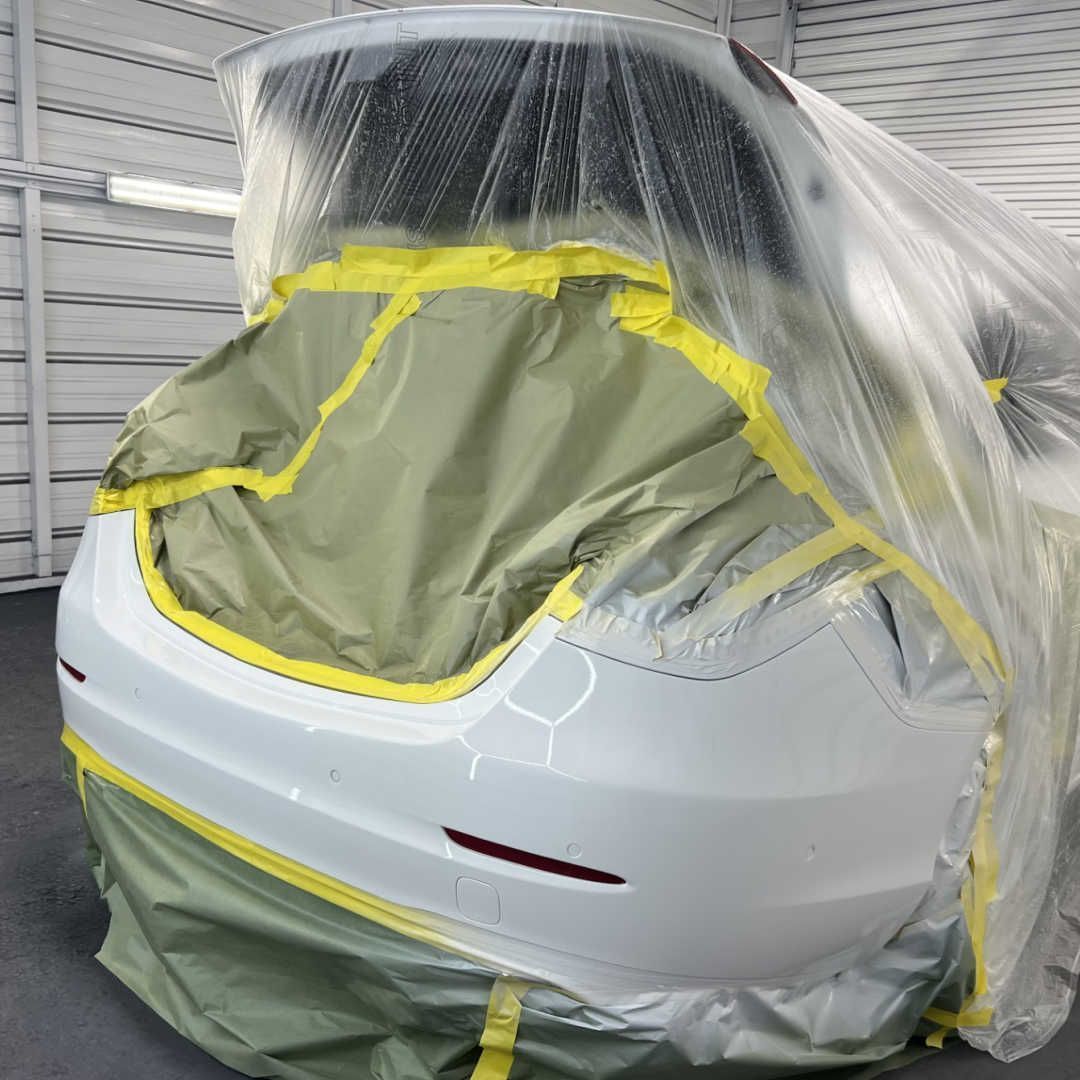 A white car bumper in an auto body shop, masked with plastic sheeting and yellow tape in preparation for painting.