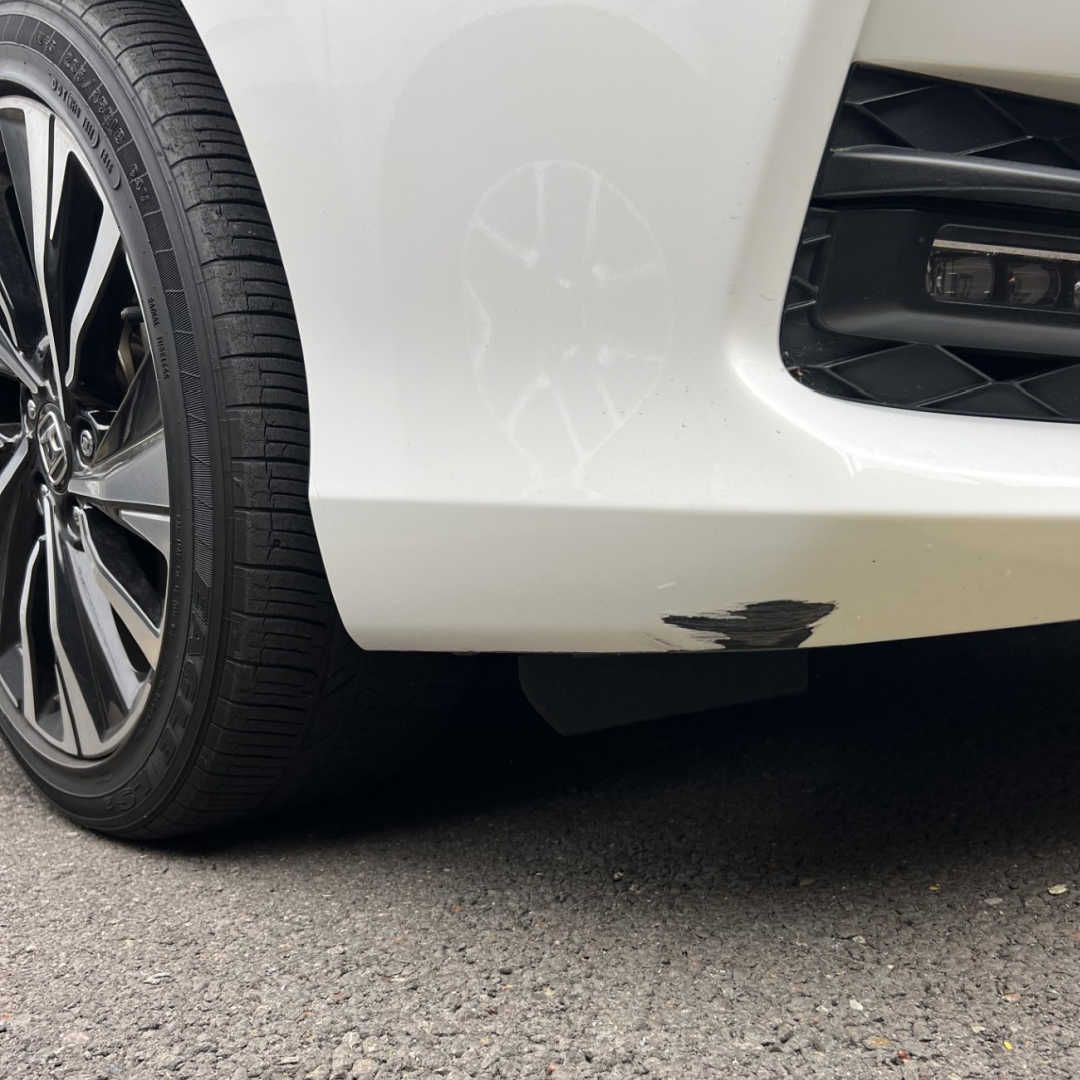 Close-up of a white car bumper showing a patch of dark paint damage near the front wheel.
