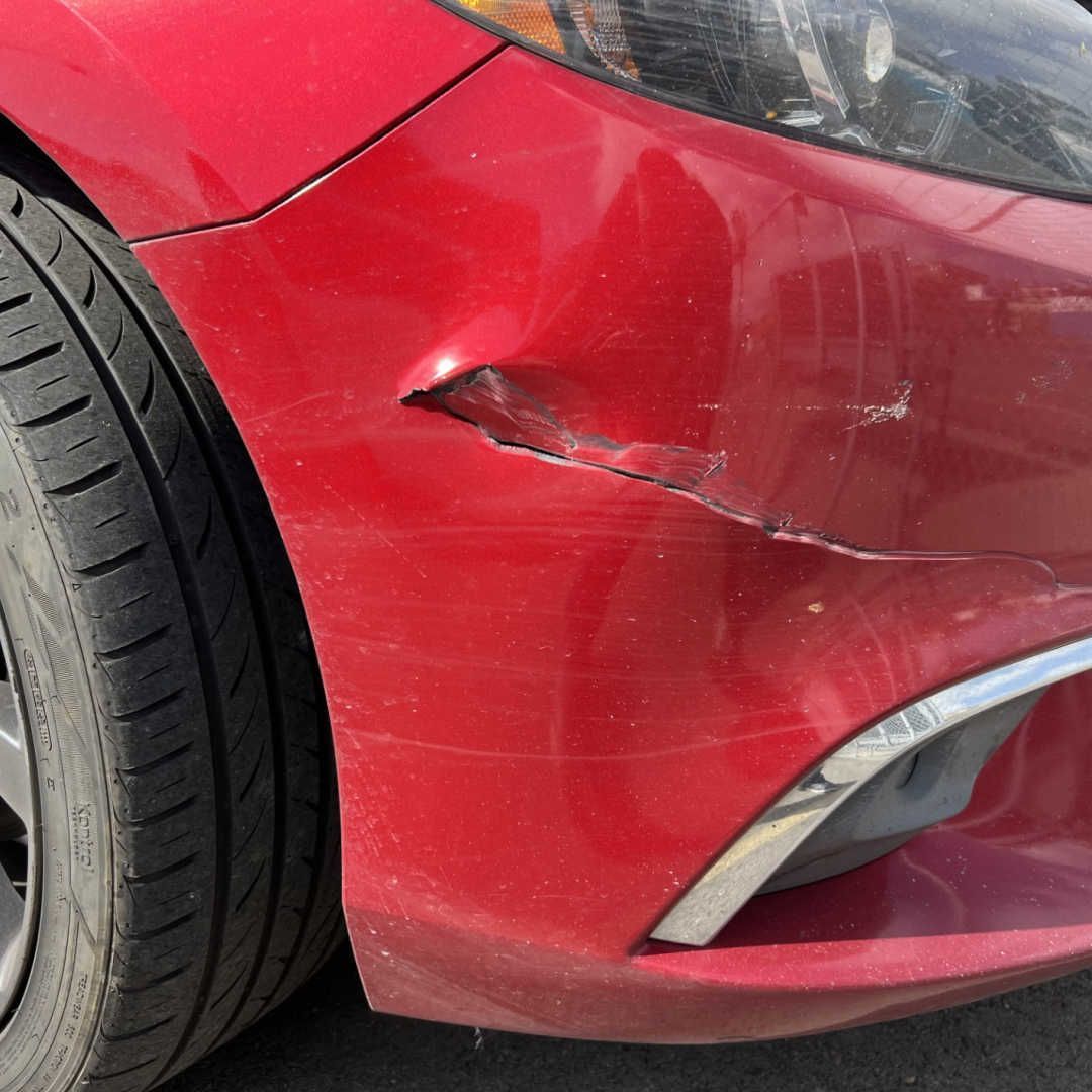 A close-up of a damaged red car bumper with a significant dent and a crack near the front wheel.
