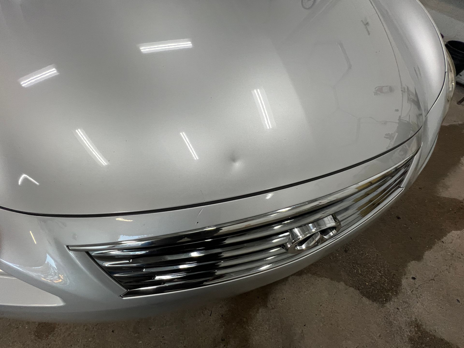 A silver car hood featuring a small, circular dent located near the front grille.