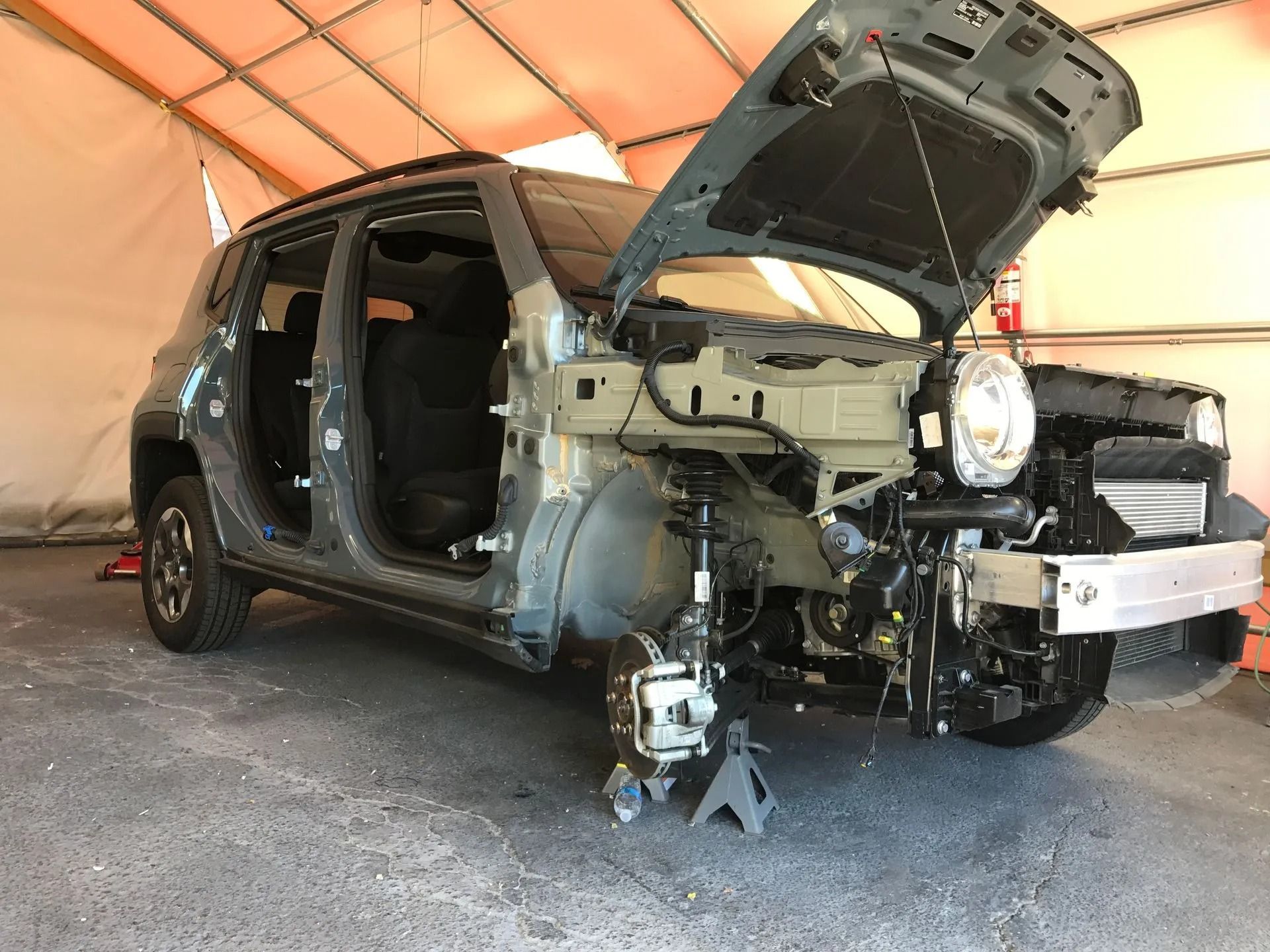 A gray car chassis missing its front fenders and doors, supported by jack stands inside a covered structure.