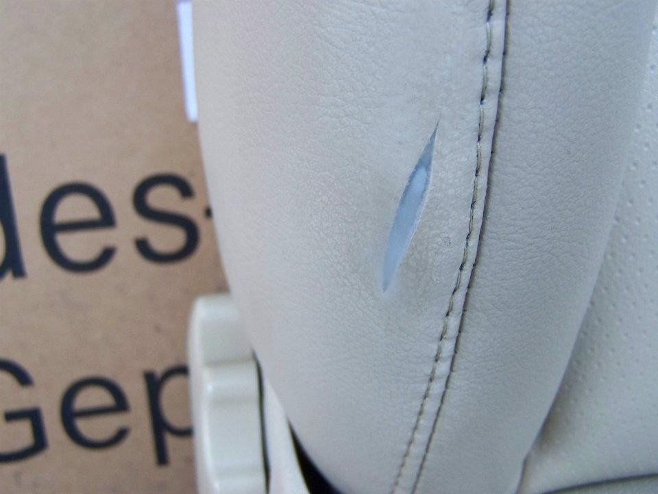 A close-up of a rip in the beige synthetic leather upholstery of a car seat, positioned near a stitched seam.