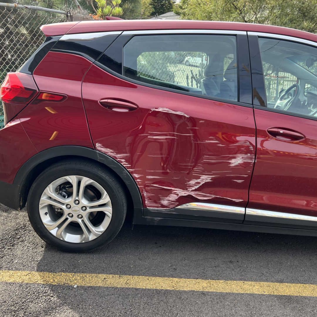 A red car parked on asphalt with significant damage and deep dents on the rear passenger-side door.