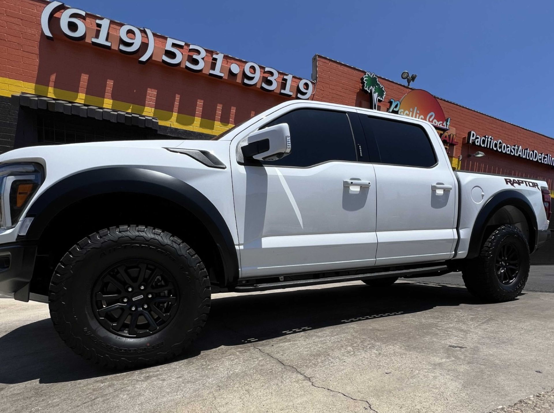 A white Ford F-150 Raptor pickup truck parked in front of a brick building with a phone number on the wall.