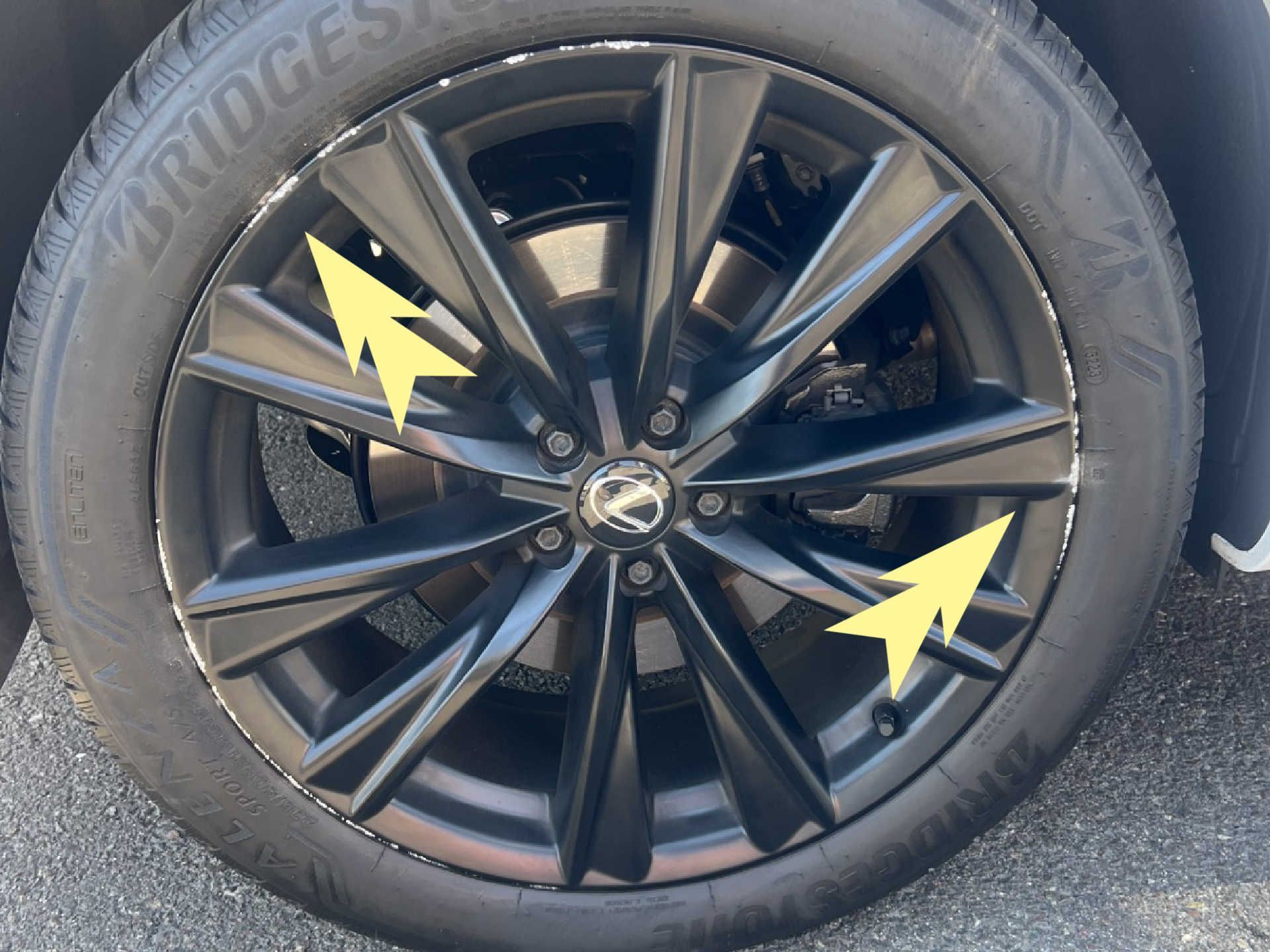 Black Lexus alloy wheel with yellow arrows pointing to signs of curb rash on the rim edge.
