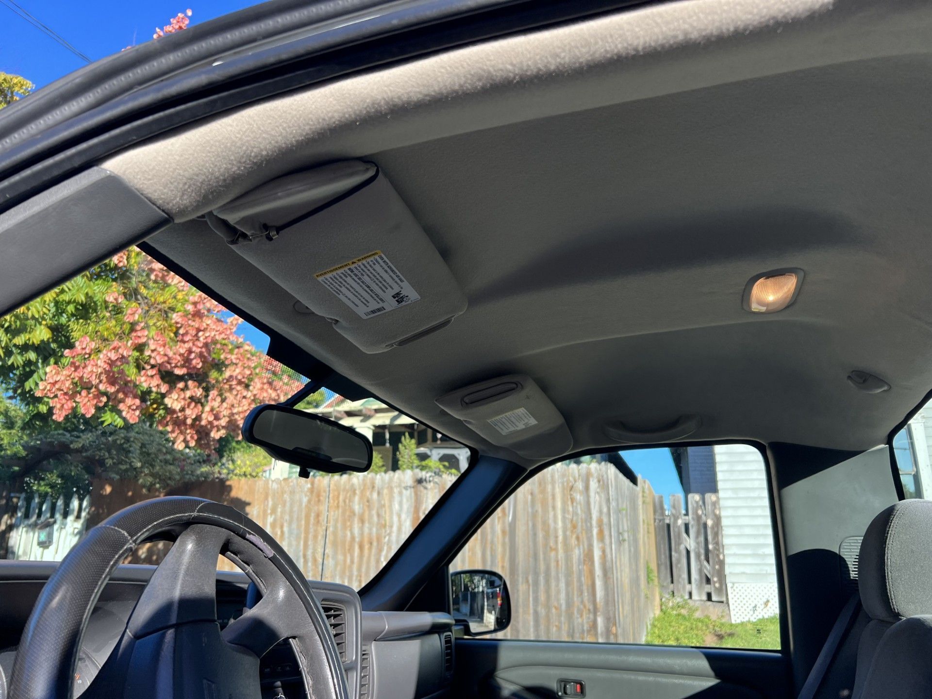 Interior of a vehicle cabin looking out through the windshield at a sunny suburban street with trees and a wooden fence.