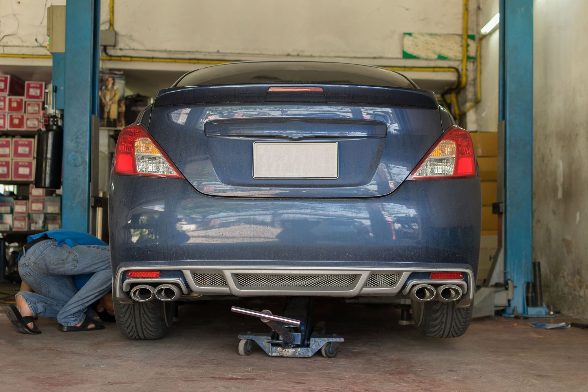Blue car lifted in a garage for repair, with a mechanic working near the rear wheel