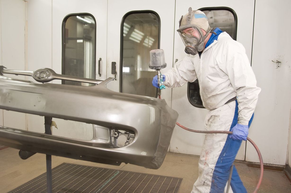 Worker in white protective suit spray-painting a car bumper in a paint booth.