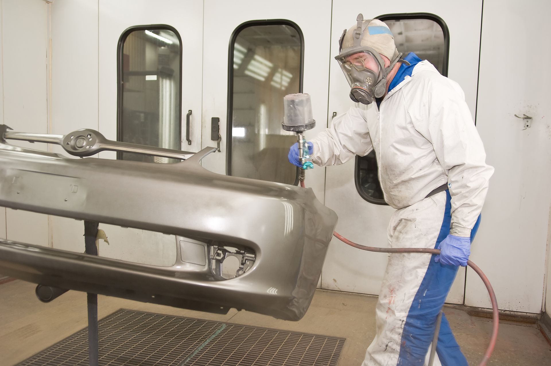 Worker in white protective suit spray-painting a car bumper in a paint booth.