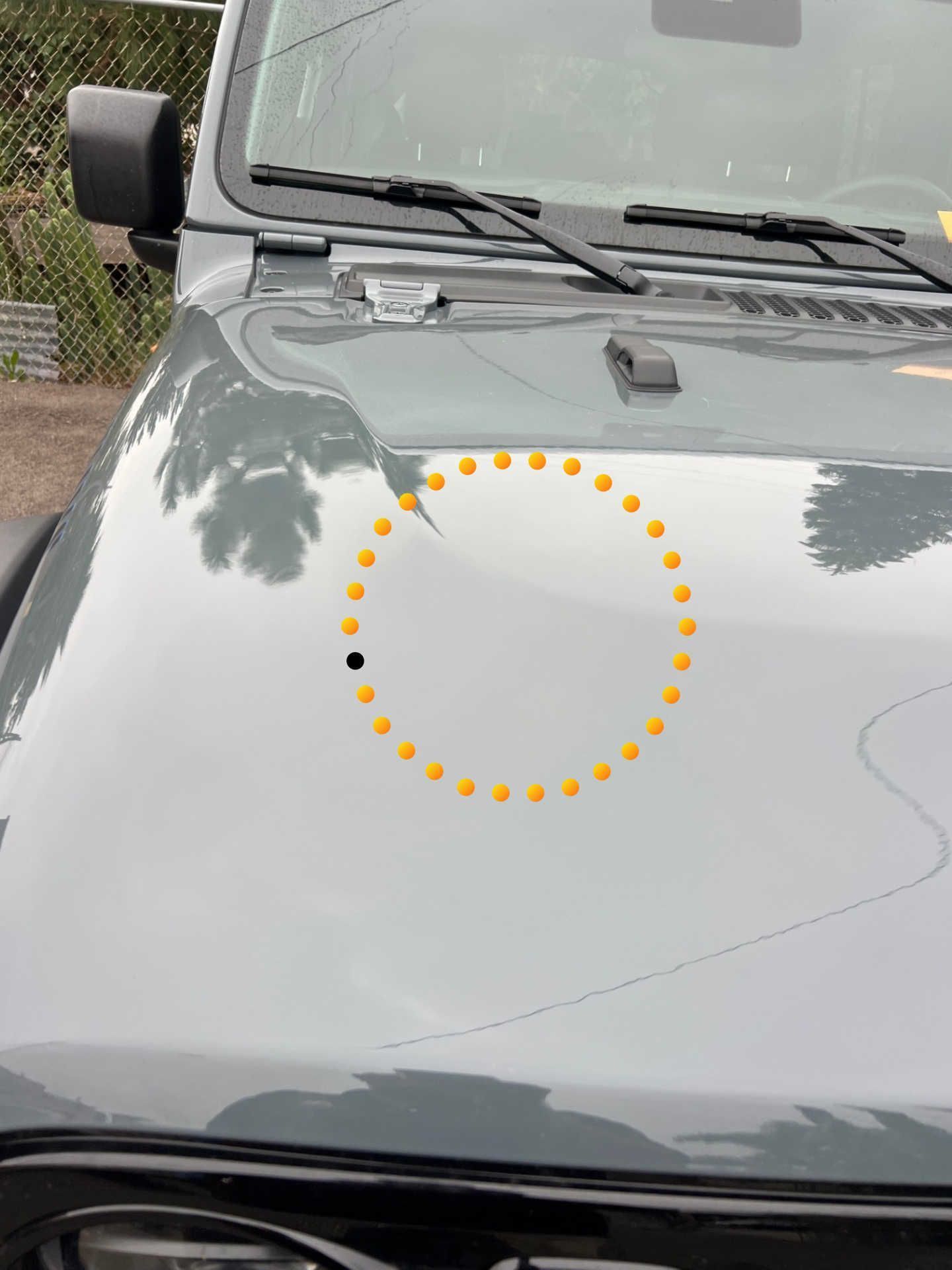 A close-up of a gray car hood featuring a small dent highlighted by a circular graphic overlay and a black dot marker.