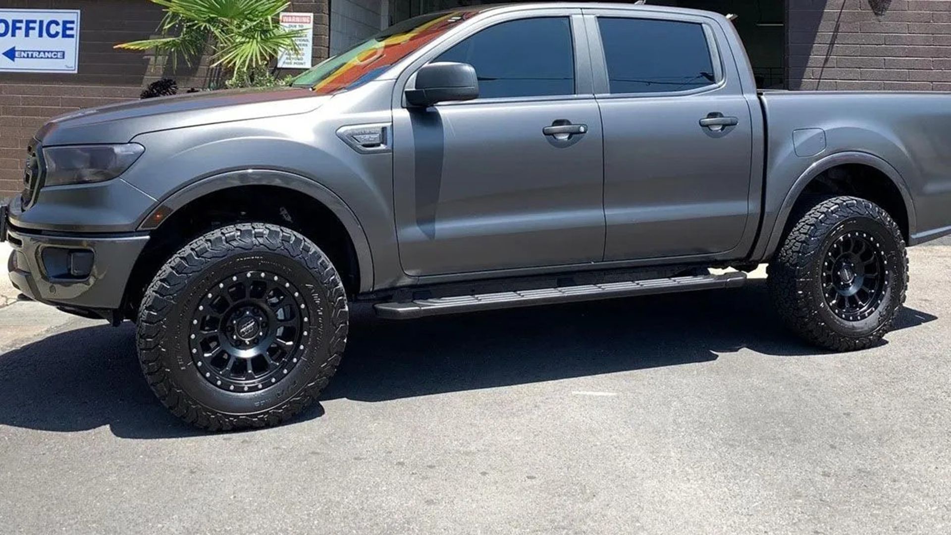 A dark gray lifted Ford Ranger pickup truck parked on an asphalt lot in front of a building office sign.