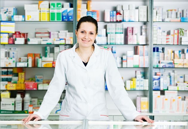 Pharmacist in a white coat standing behind a counter in a pharmacy, with medicine shelves in the background