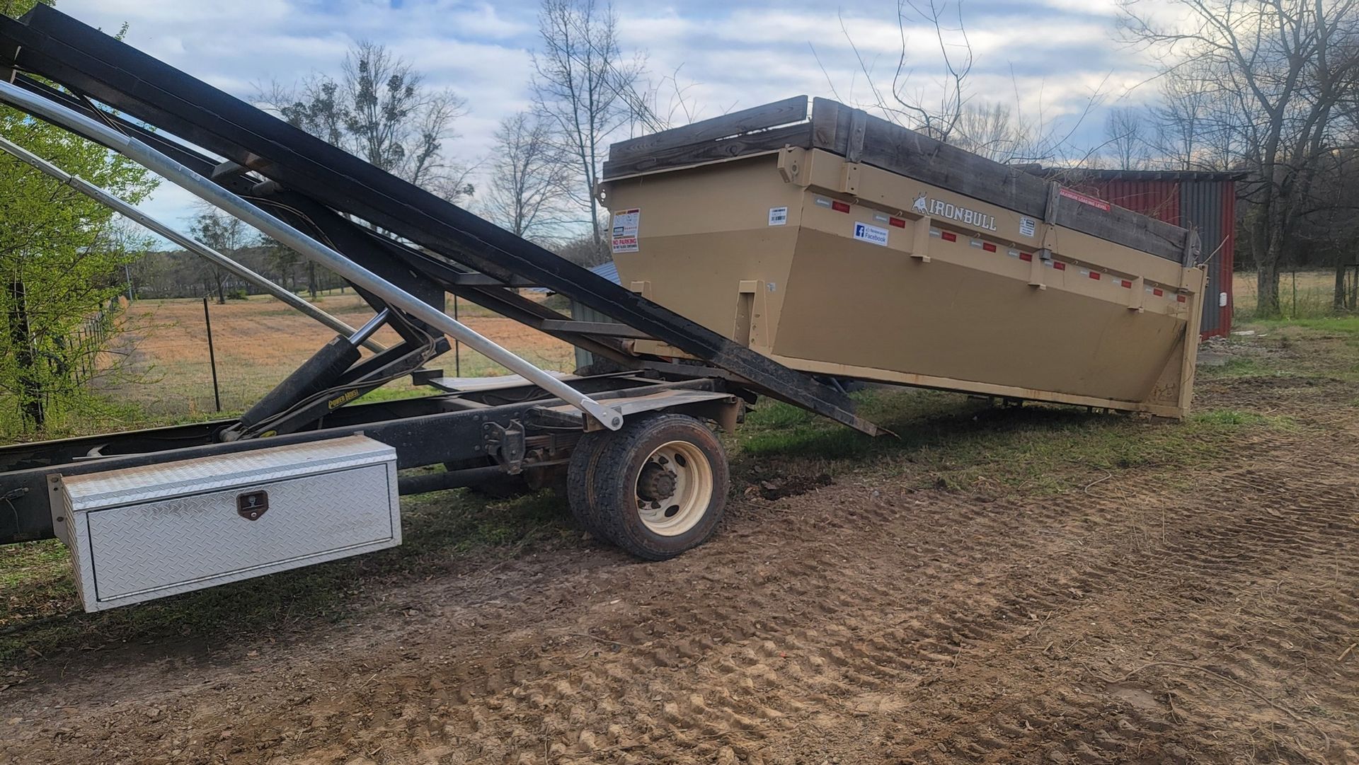 A dumpster is being pulled by a dump truck in a dirt field.