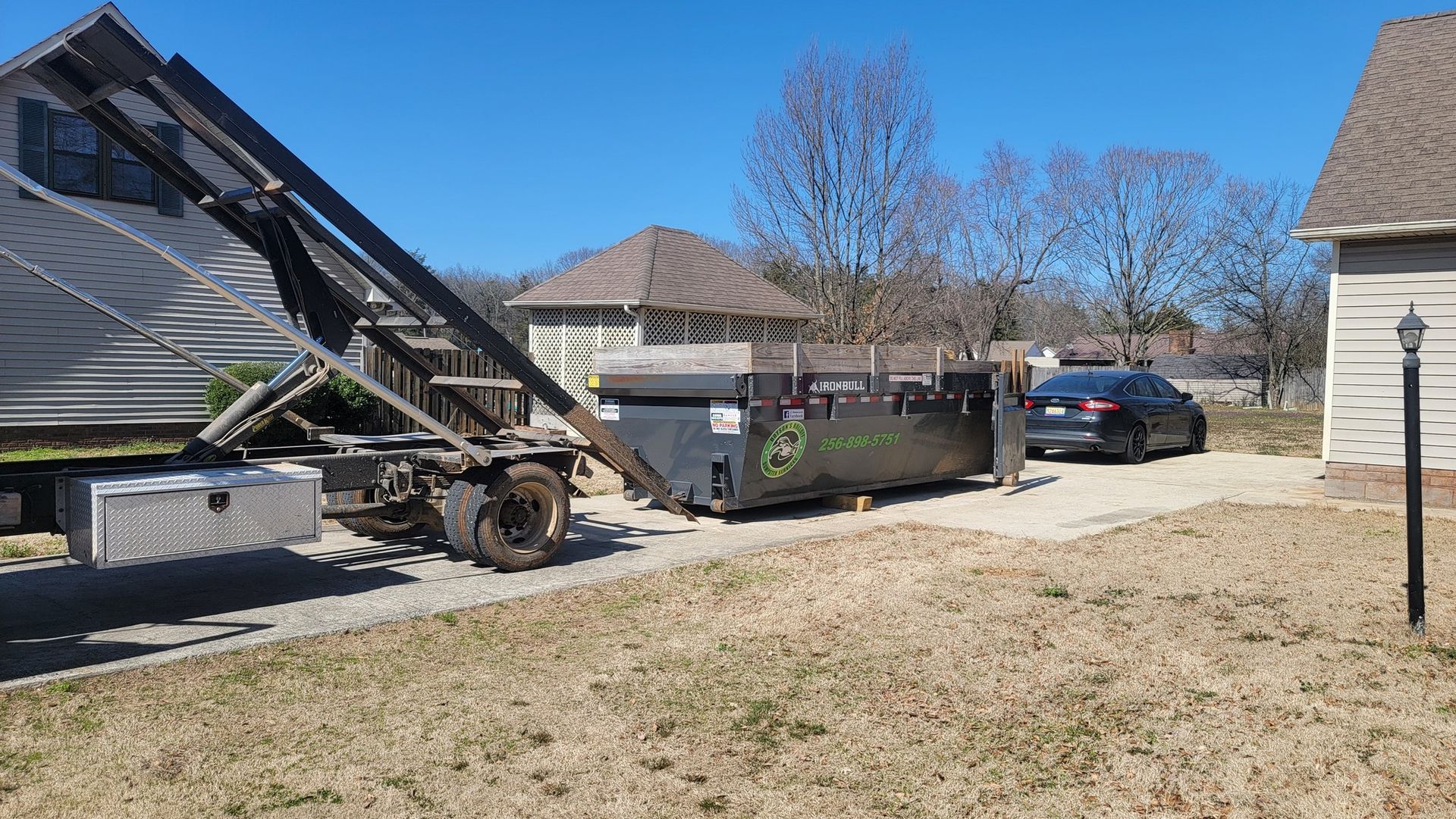A dumpster is being loaded into a truck in a driveway next to a house.