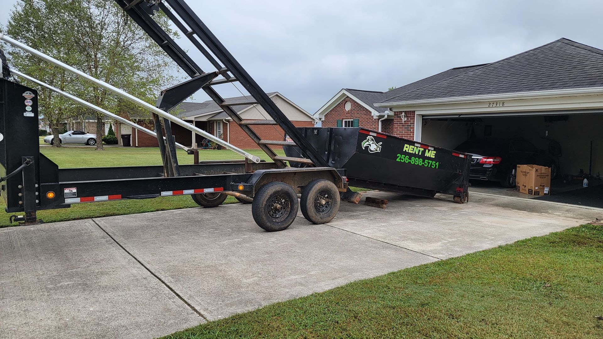 A dumpster is sitting on a trailer in a driveway next to a garage.