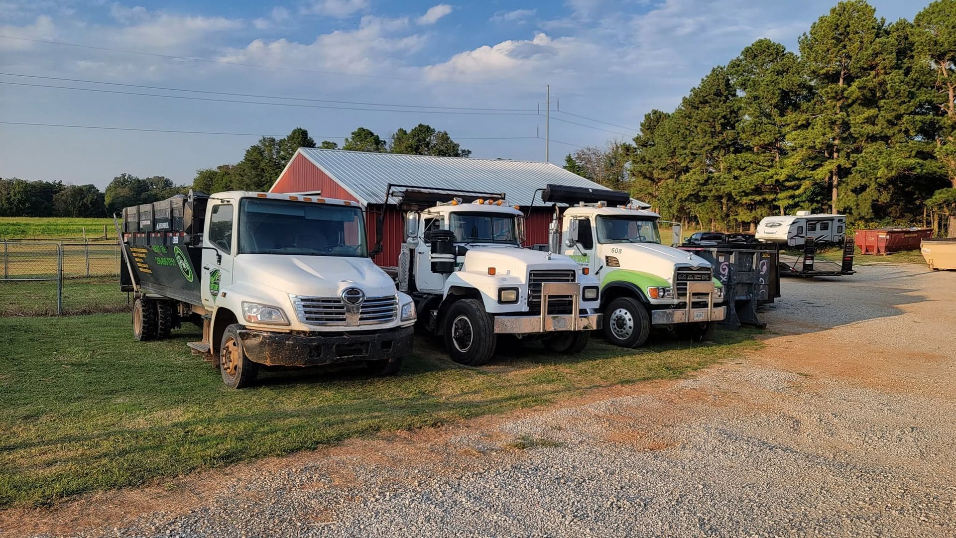 Three utility trucks parked on gravel beside a barn in a grassy rural yard