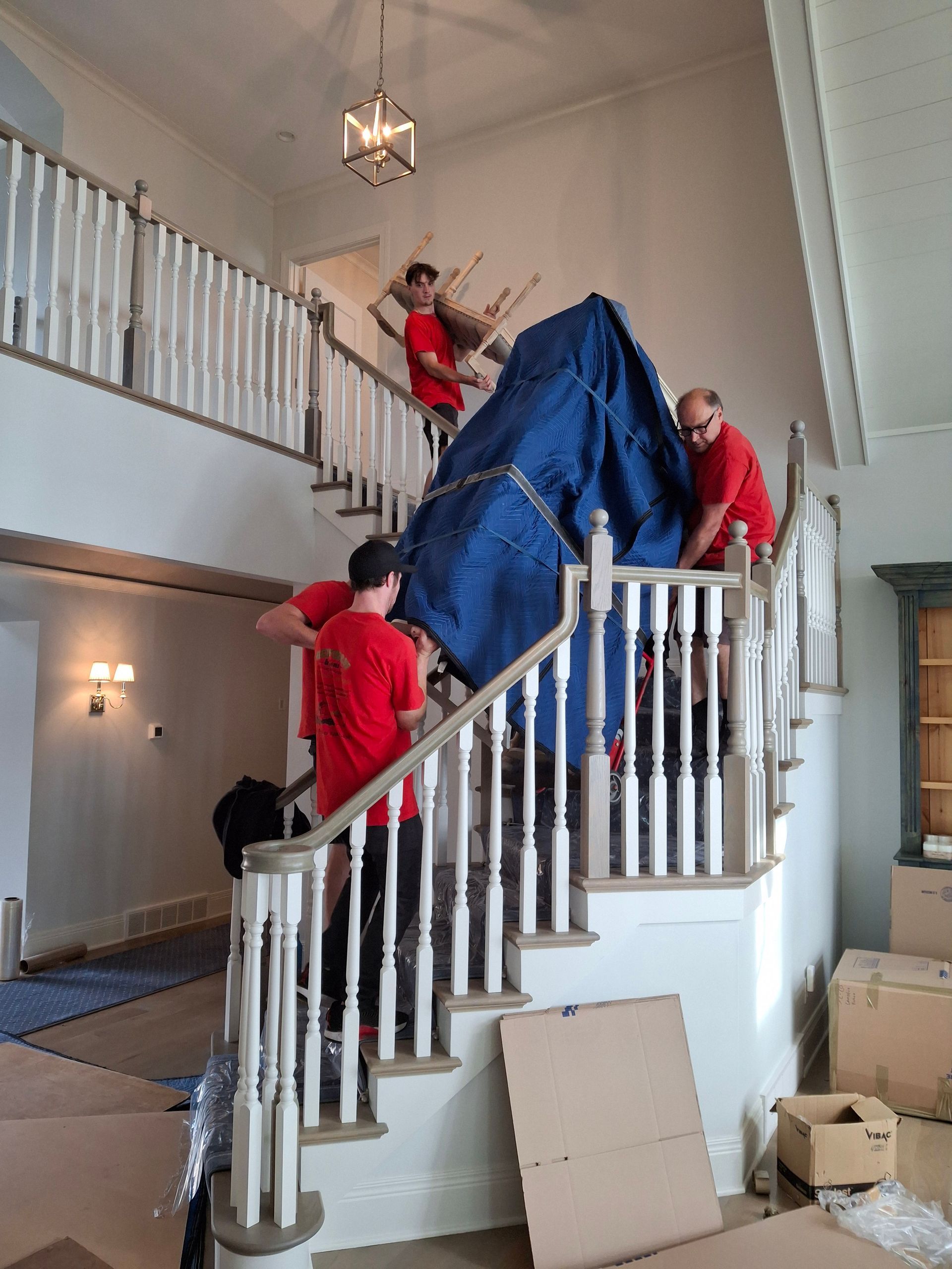 Movers carrying a large, blue-covered object up a staircase with white balusters.