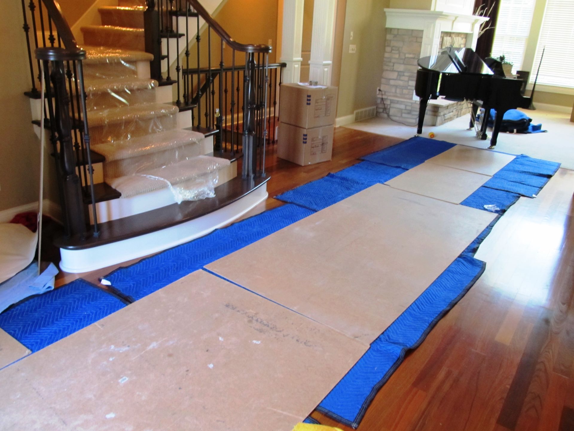 Cardboard and blue moving pads protect wood floor. Staircase with plastic wrap, piano in background.