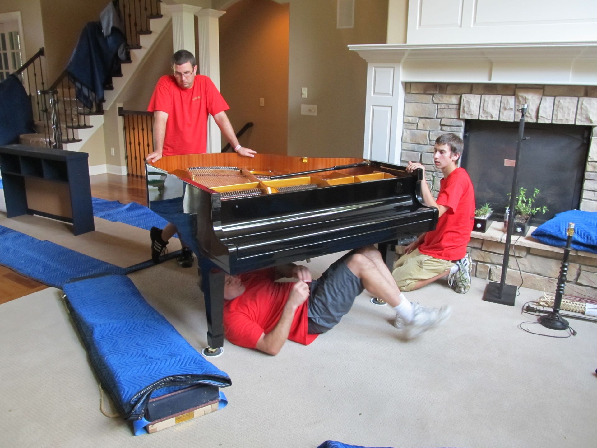 Three people in red shirts lifting a black piano in a home; blue moving pads are on the floor.
