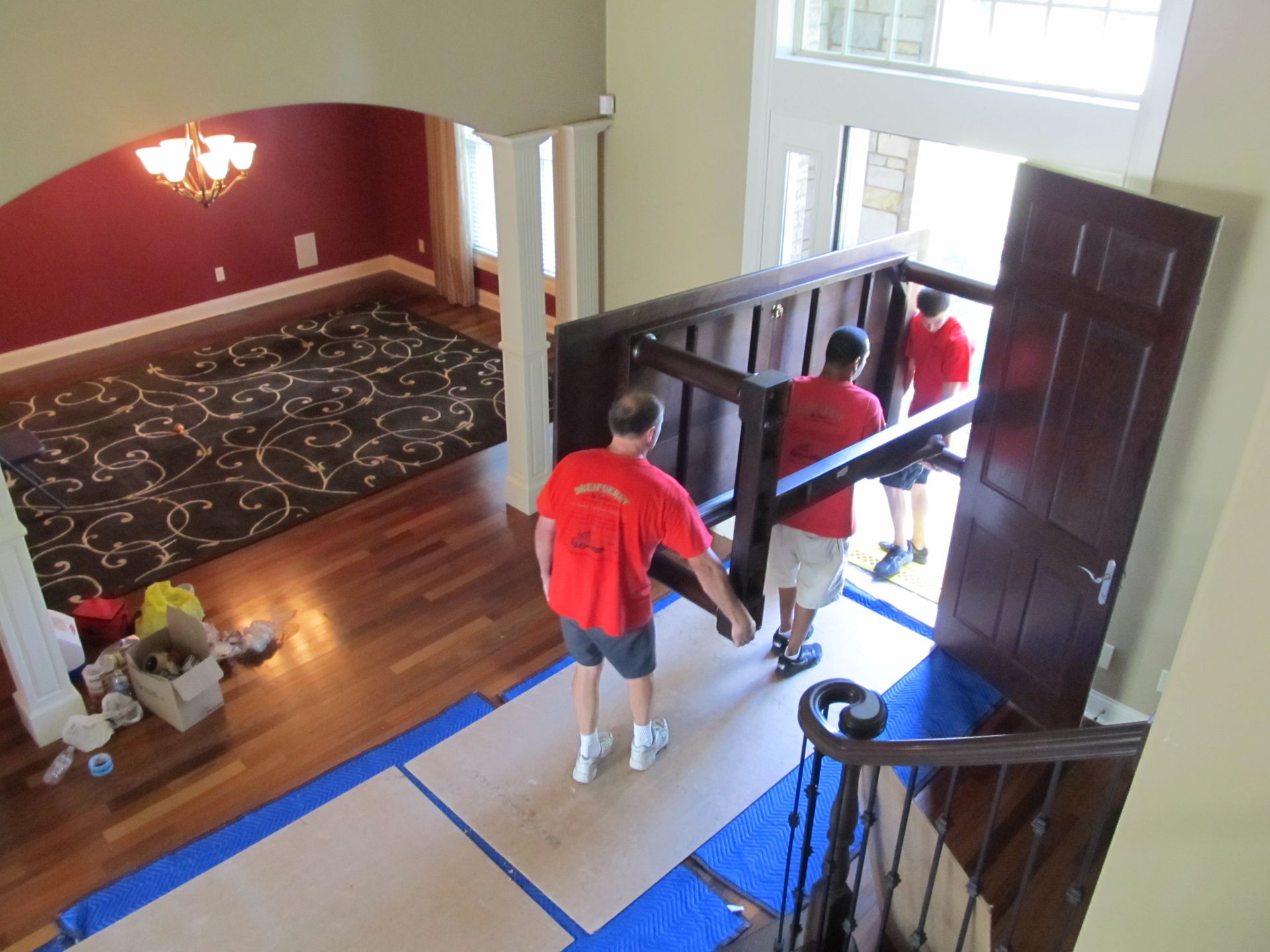 Movers carrying a large wooden headboard through a doorway into a house with hardwood floors.