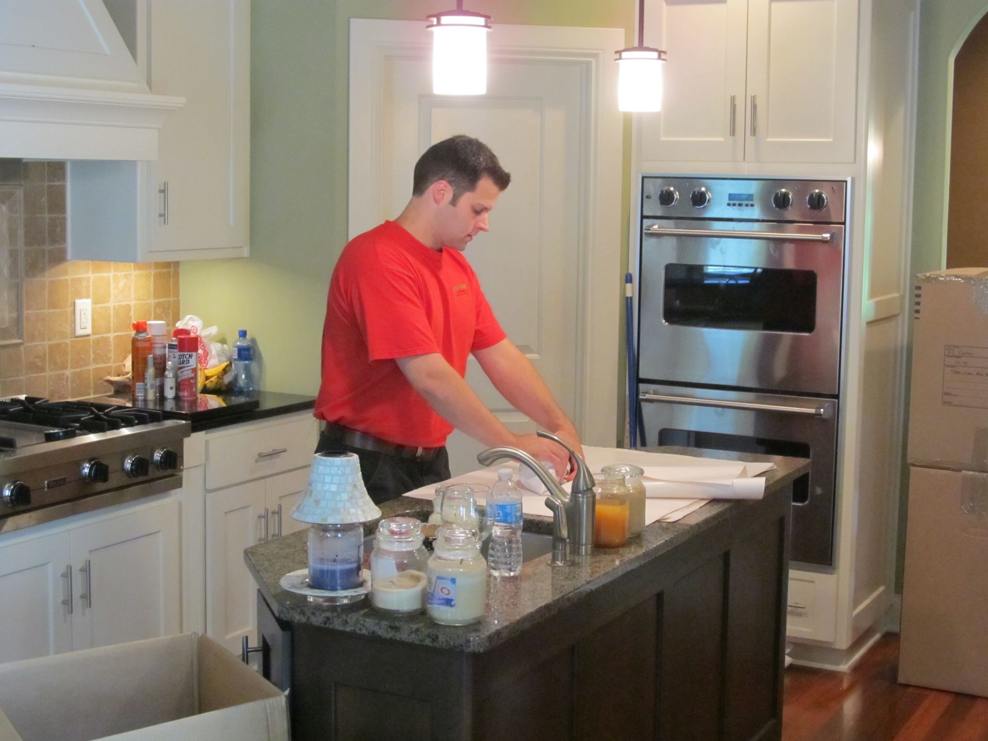Man in red shirt in a kitchen, looking at papers on a counter. White cabinets, stainless steel oven, and boxes are nearby.