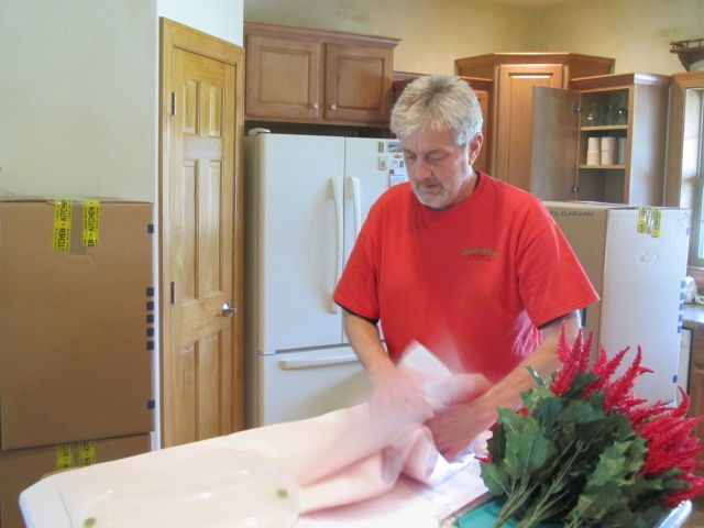 Man in red shirt wrapping something on a table, with boxes, cabinets, and a refrigerator in the background.