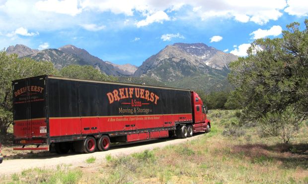 Red semi-truck with black trailer, "DREIFUERST" logo, on dirt road with mountains and blue sky in background.