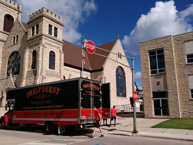A red moving truck parked on a street in front of a stone church and modern building.