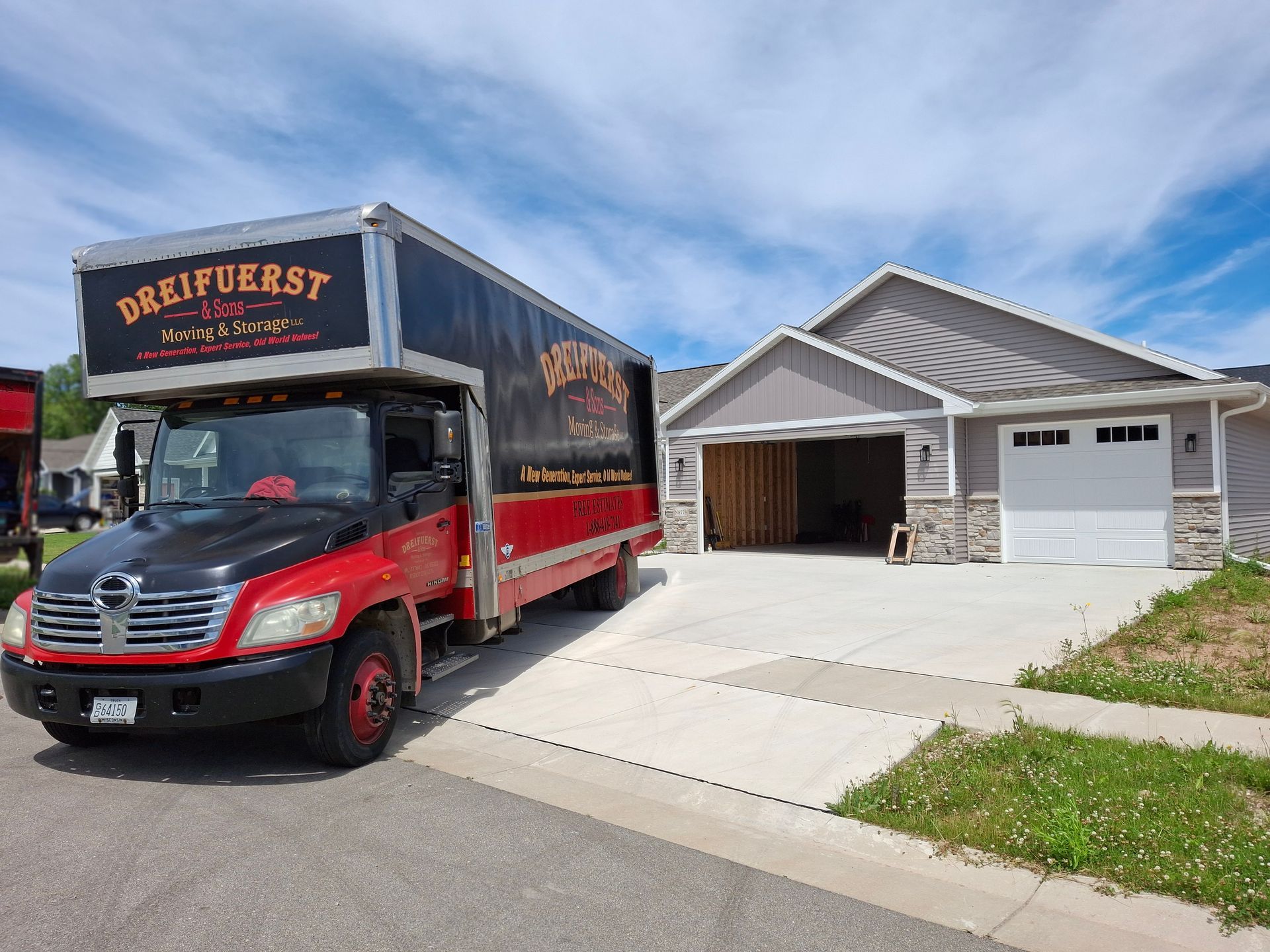 Red and black moving truck parked in front of a gray house with open garage. Sunny day.