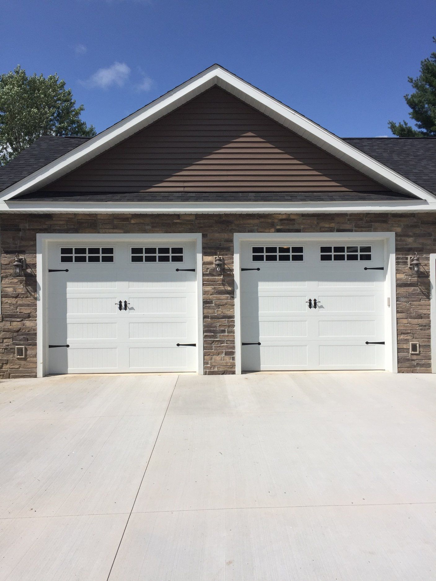Two white garage doors with black accents, set in a stone facade, under a brown-sided gable roof.