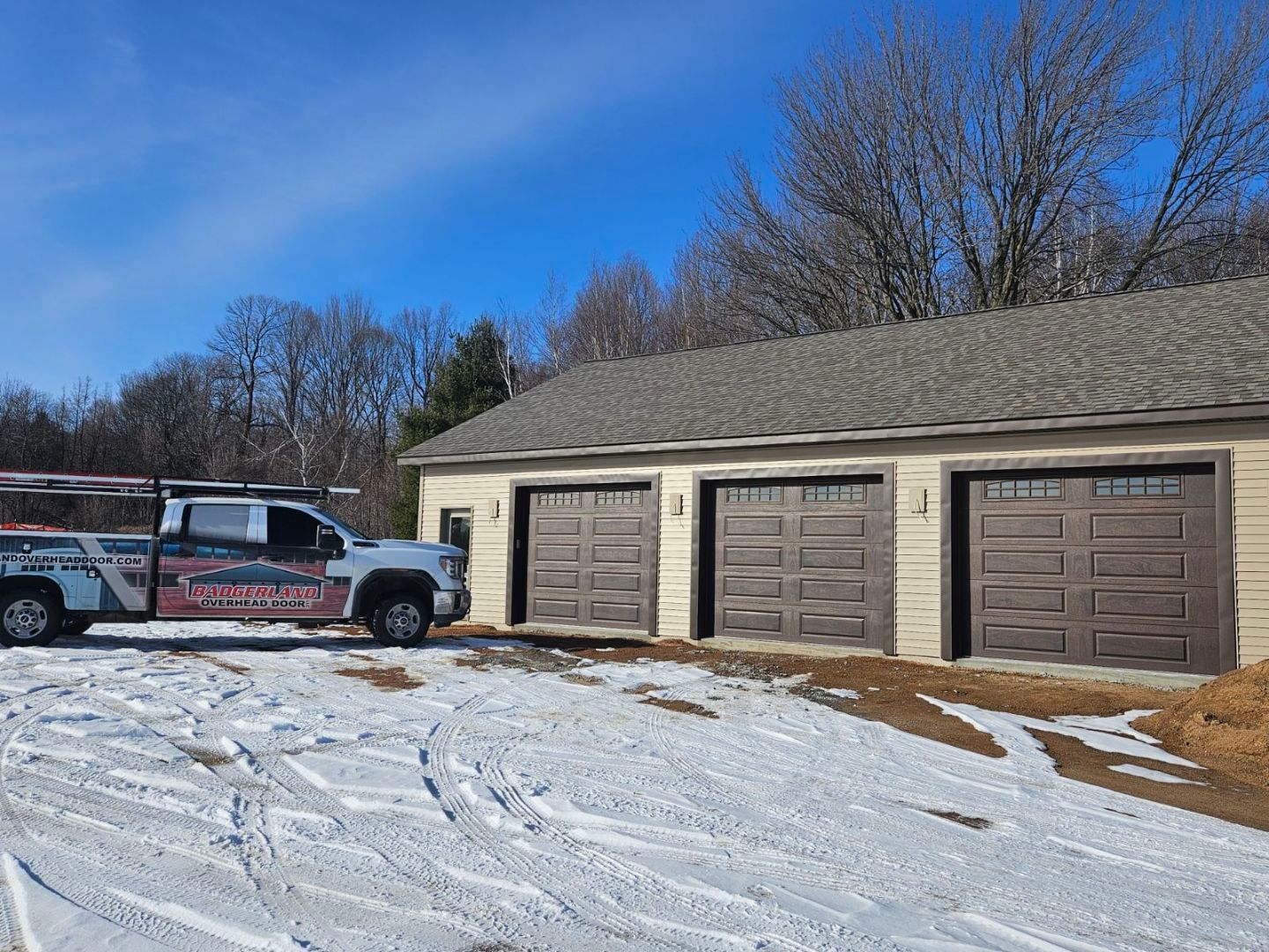 Truck parked near a three-car garage in a snowy landscape under a blue sky.