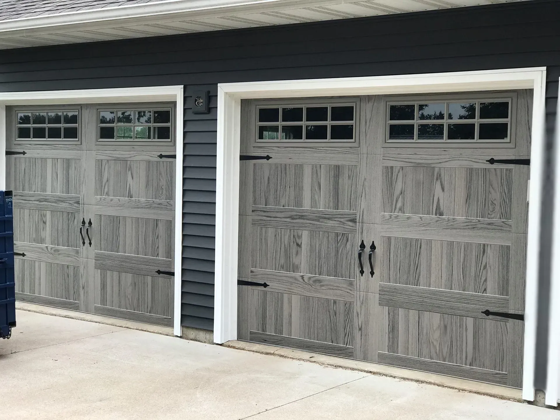 Two gray wooden garage doors with windows and black hardware on a house with dark blue siding.