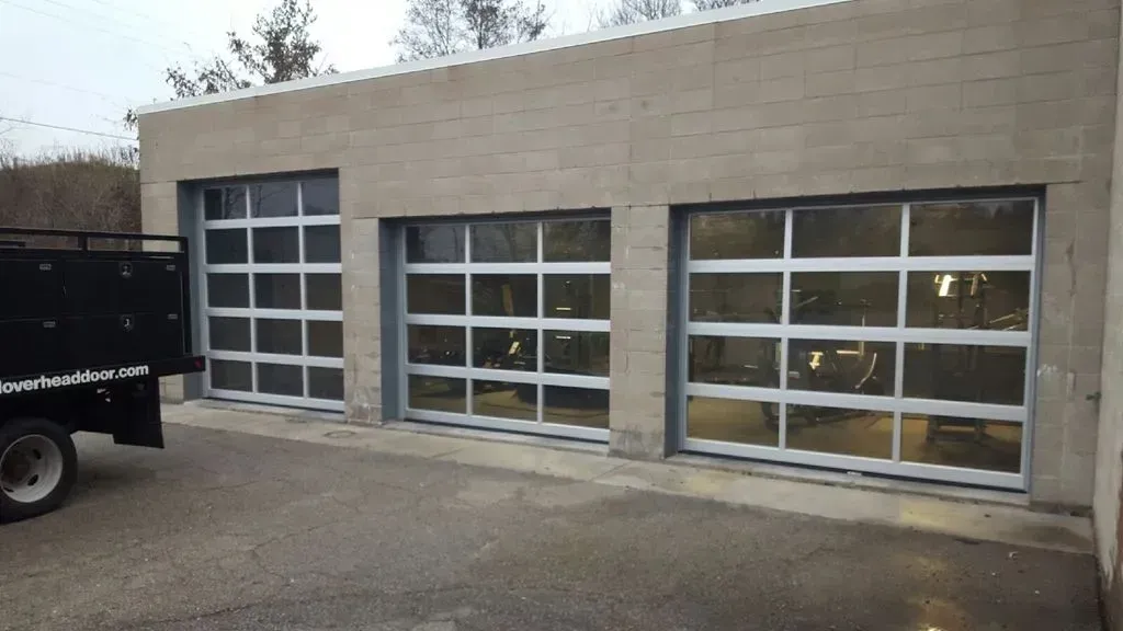 Three garage doors with glass panels on a concrete building. A truck is parked to the left.