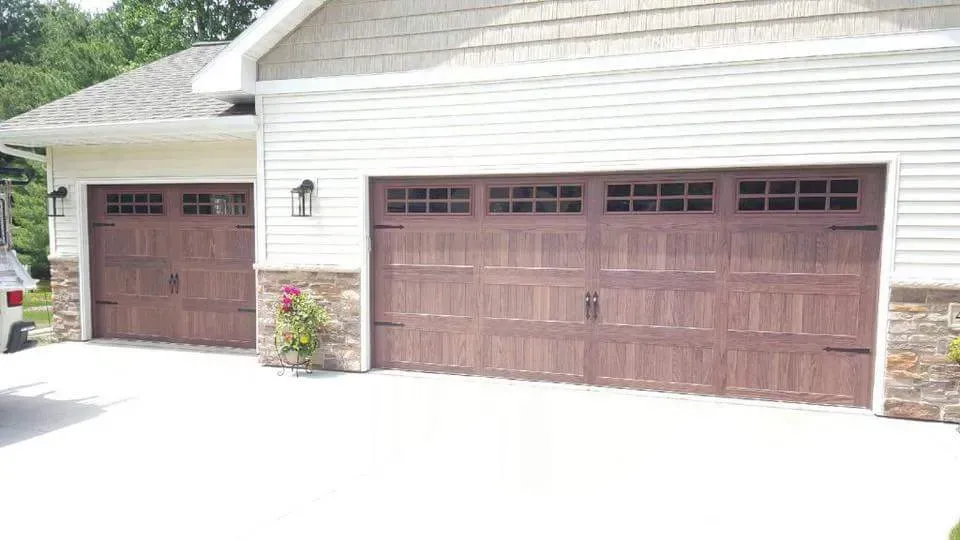 Two brown wooden garage doors on a house with off-white siding and a concrete driveway.
