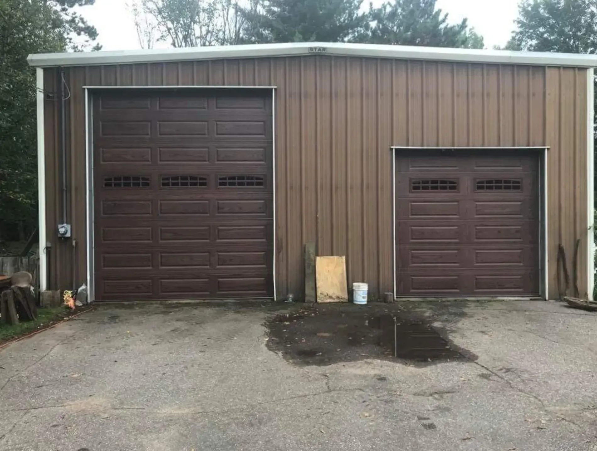 Brown metal garage with two doors on a gray concrete surface; puddles in front.
