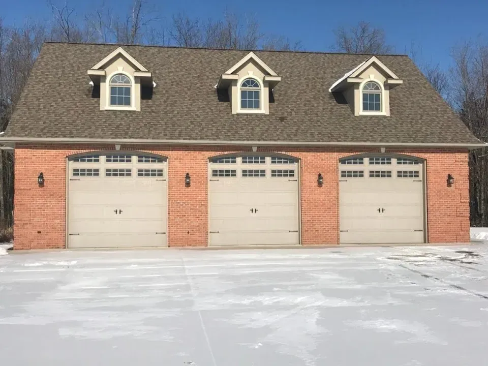 Three-car garage with tan doors, brick facade, and dormers under a brown roof. Snowy driveway.