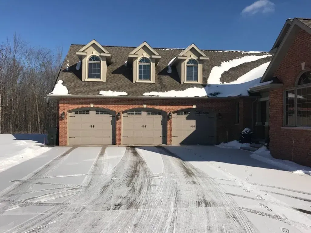 Three-car garage with snow-covered roof and driveway, brick exterior, and dormer windows under a blue sky.