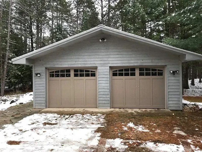 Two-car garage with tan doors and gray siding, snow on ground.