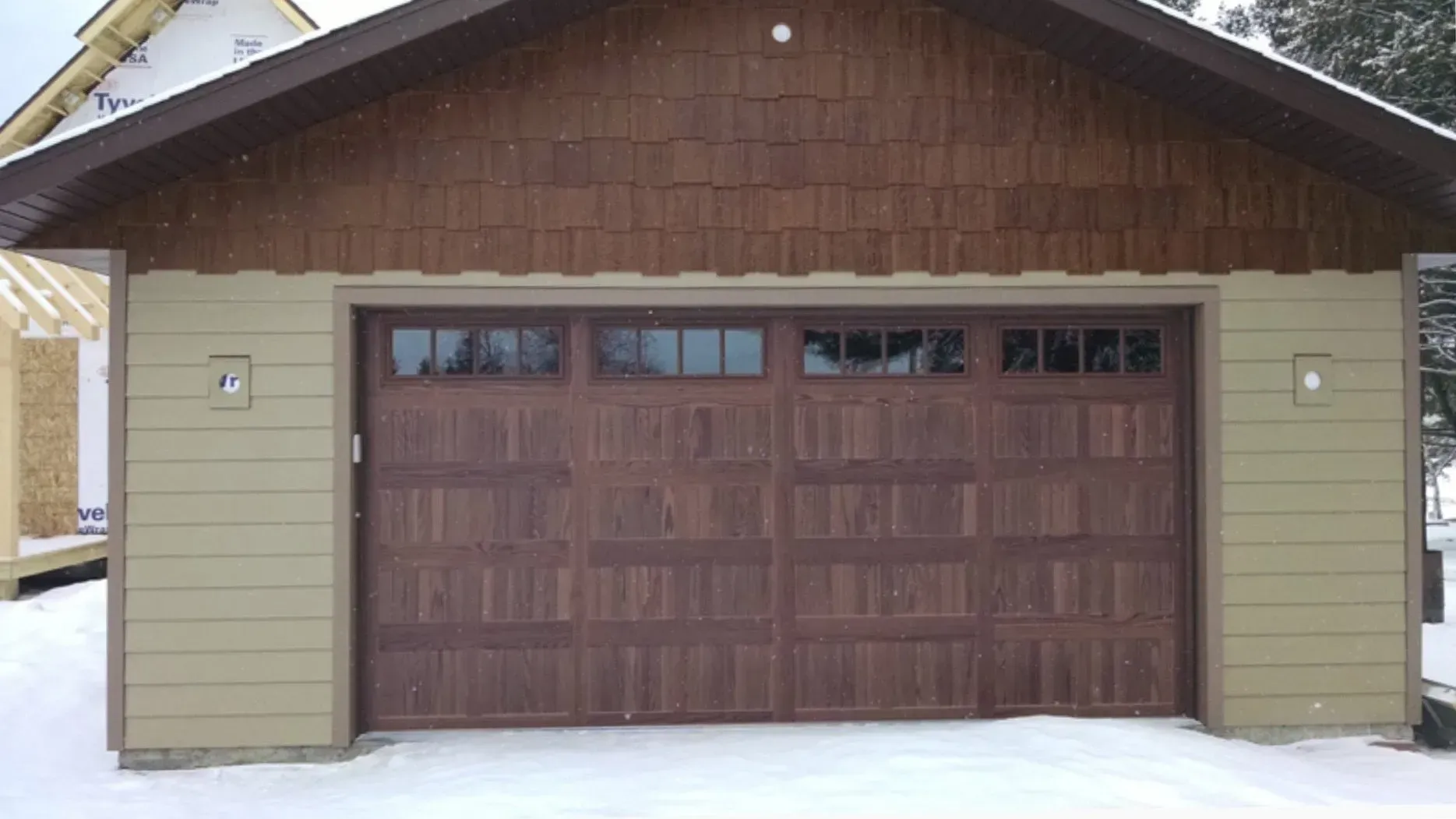 Brown garage door with windows, under a brown shingled gable, beige siding, snow.