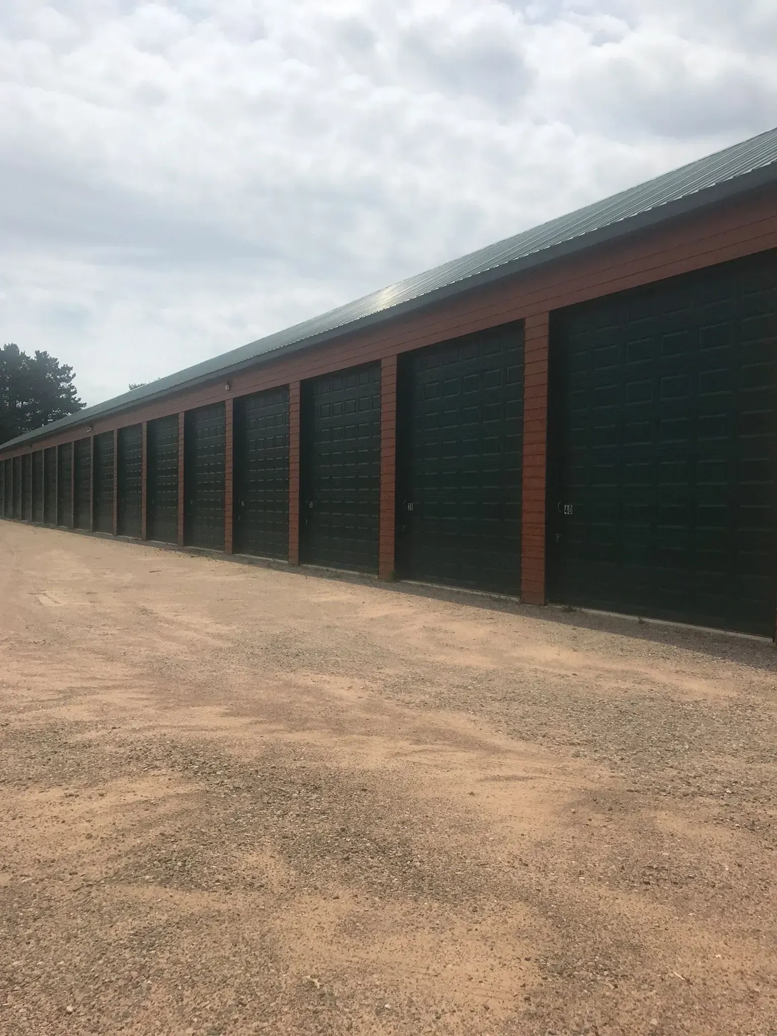 Row of green storage unit doors along a gravel drive, under a cloudy sky.