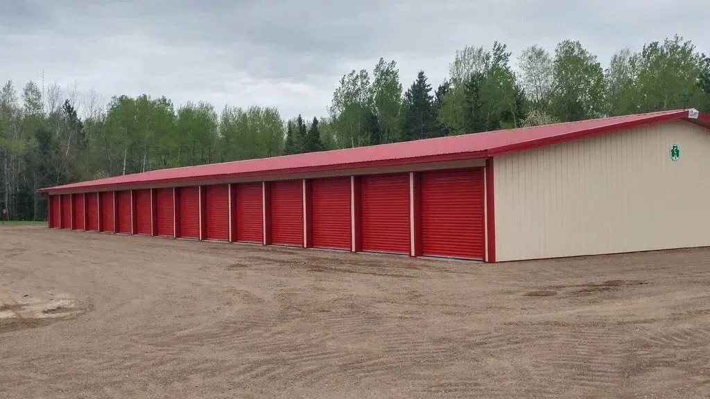 Red-doored storage units in a long, beige building under a red roof, located outdoors on a gravel lot.