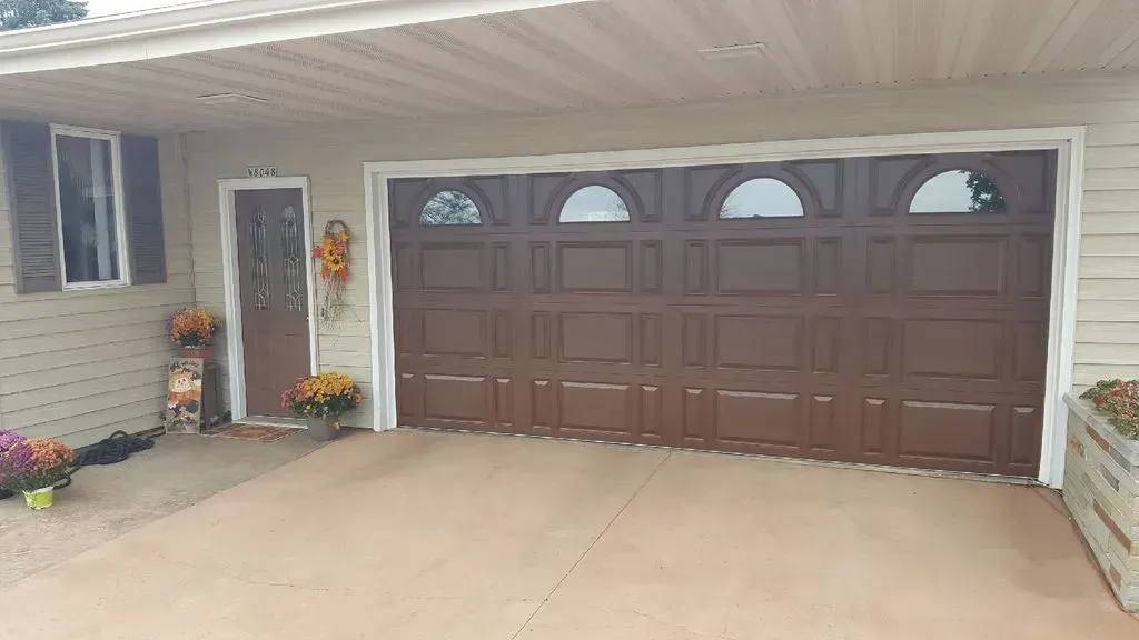 Brown garage door with arched windows and a brown front door. Beige siding with colorful autumn flowers.