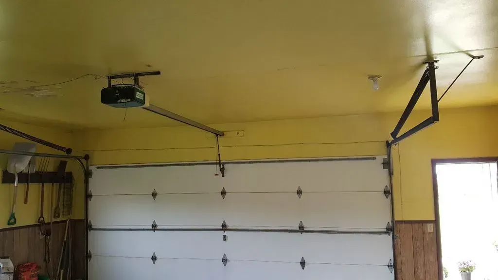 A white garage door is open, inside a garage with yellow ceiling and opener.