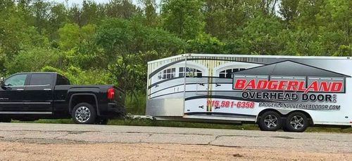 A black pickup truck towing a trailer with "Badgerland Overhead Door" advertising on it, near green trees.