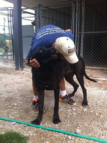 A man wearing a blue shirt that says texas on it is hugging a black dog