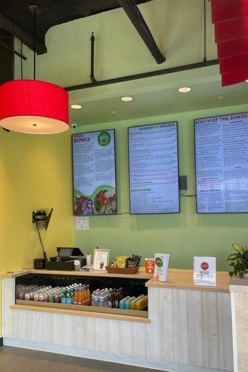 A light green shop counter with a red hanging lamp, a refrigerated drink display, and three menu boards on the wall.
